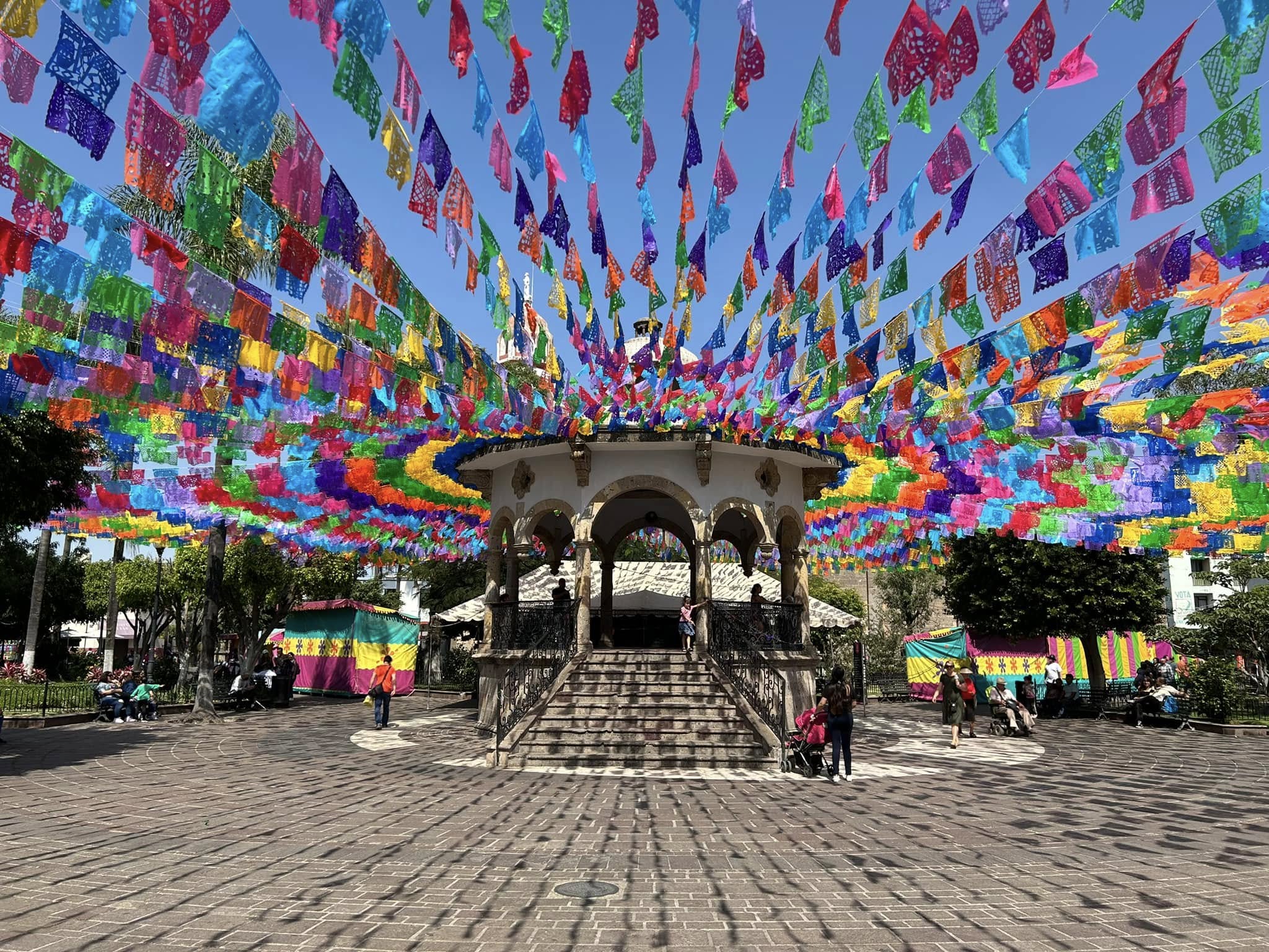The stone kiosco in the main plaza of Ajijic under a clear blue sky, with hundreds of strings of brightly colored papel picado stretched overhead and their shadows falling across the brick pavement